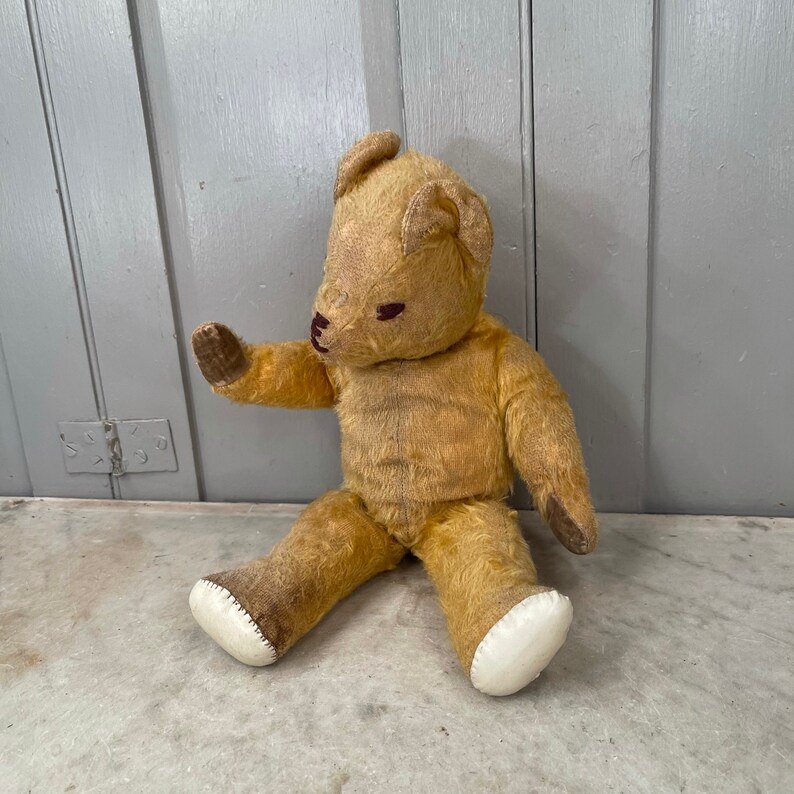 Puede incluir: Un oso de peluche vintage de color beige con ojos marrones y una nariz cosida. El oso est&aacute; sentado en una superficie de color claro con los brazos extendidos.