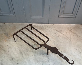 May include: A rusty metal fireplace tool with a long handle and four prongs. The tool is designed to lift and move logs in a fireplace.