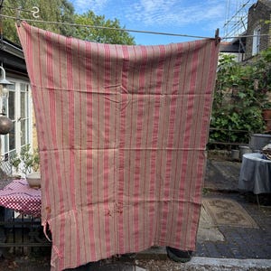 May include: A large, rectangular, vintage textile with vertical red stripes on a tan background, hanging on a clothesline. The fabric appears worn and weathered, with visible imperfections. The textile is suspended outdoors, suggesting it is drying in the sun.