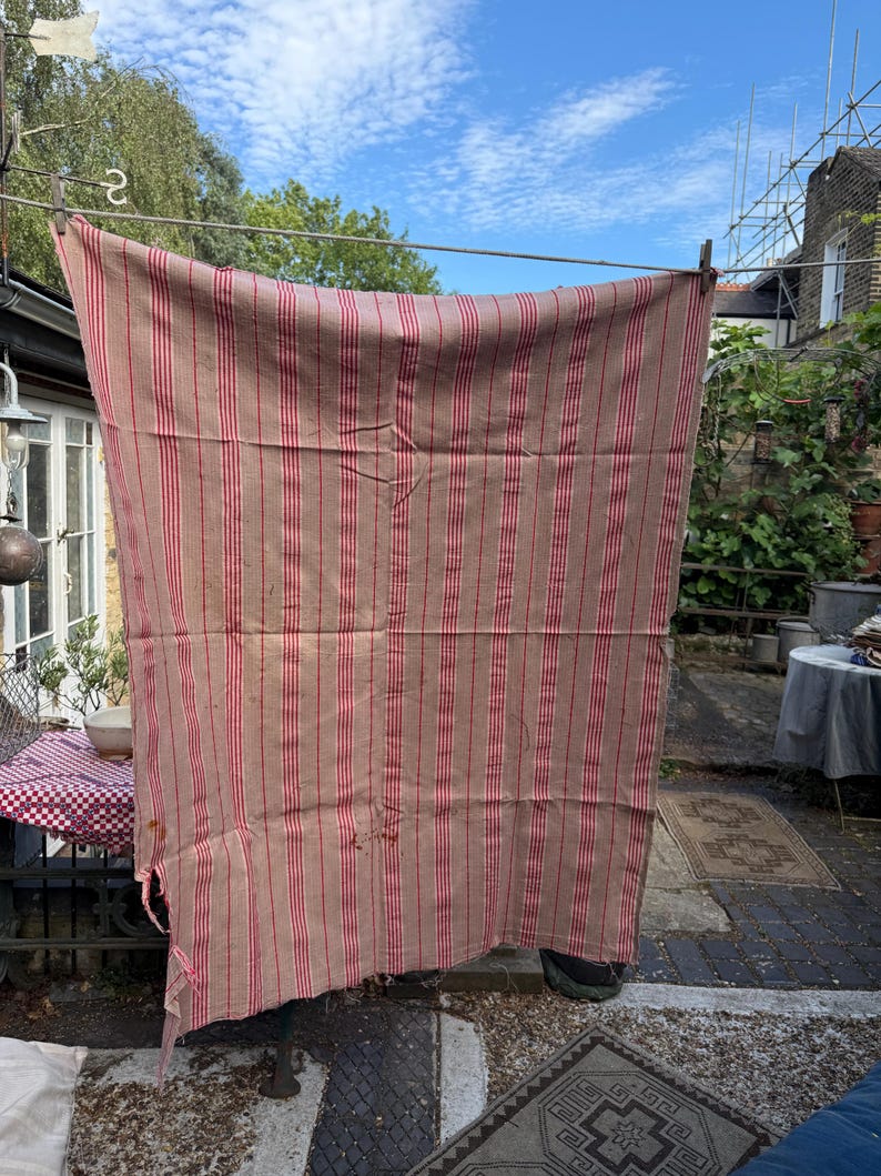 May include: A large, rectangular, vintage textile with vertical red stripes on a tan background, hanging on a clothesline. The fabric appears worn and weathered, with visible imperfections. The textile is suspended outdoors, suggesting it is drying in the sun.