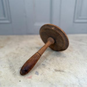 Puede incluir: Un molde de mantequilla de madera vintage con una parte superior redonda y plana y un mango largo y redondeado. La madera tiene un tono marrón cálido con vetas visibles y signos de desgaste, lo que sugiere edad y uso. El mango está unido a la parte superior con una pequeña pieza cilíndrica.