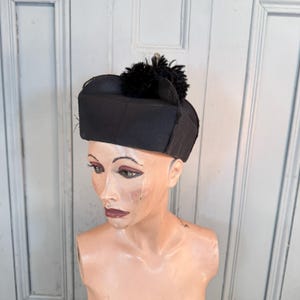 May include: A structured black hat with a flat top and a prominent black pom-pom centred on the crown. The hat features a stiff, cylindrical band design that sits horizontally, providing a clean, geometric silhouette against the mannequin head.