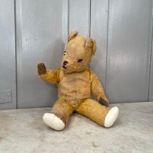 Puede incluir: Un oso de peluche vintage de color beige con ojos y nariz bordados en marr&oacute;n. El oso est&aacute; sentado en una superficie blanca con el brazo izquierdo levantado y el brazo derecho descansando en su regazo. El oso tiene tela blanca en sus pies.