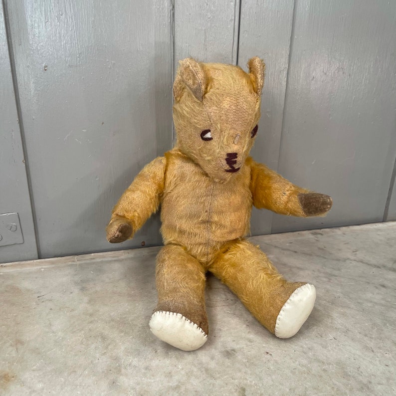 Puede incluir: Un oso de peluche vintage de color beige con patas cosidas blancas y una nariz bordada marr&oacute;n. El oso est&aacute; sentado sobre una superficie de m&aacute;rmol blanco.