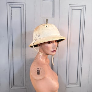 May include: A cream-colored pith helmet featuring a rounded crown, a wide brim, and a chin strap. The exterior surface shows signs of wear with dark spots and discoloration. The helmet is displayed on a mannequin head, showcasing its structure.