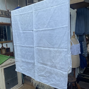 Puede incluir: Un mantel de lino blanco con un sutil patrón en relieve cuelga de un tendedero. El mantel está doblado por la mitad y parece nuevo y sin usar.