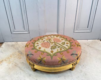 May include: A round footstool featuring a pink needlepoint top with a central floral motif of roses and green leaves. The base is constructed from gold-painted wood with carved detailing and rests on four small, low-profile feet.