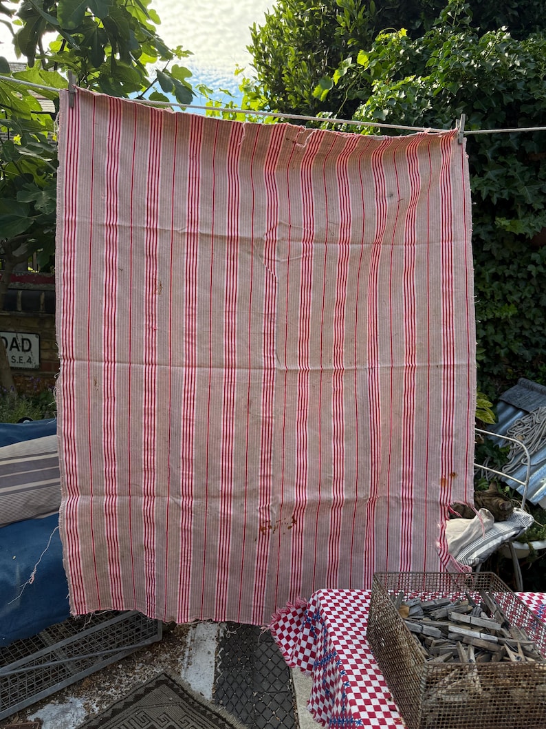 May include: A large, rectangular, vintage textile with vertical red stripes on a tan background. The fabric is hanging on a clothesline with clothespins. The textile appears worn, with some stains and imperfections.