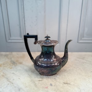 Puede incluir: Tetera antigua plateada con asa negra y boquilla curva. La tetera tiene un cuerpo bulboso y una tapa con un pequeño adorno decorativo. El metal tiene un aspecto deslustrado, mostrando toques de verde y azul.
