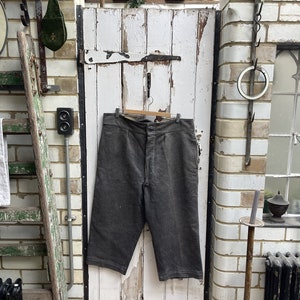 May include: A pair of dark grey striped linen trousers hanging on a wooden hanger against a white wooden door. The trousers have a button fly and are cropped just above the knee.