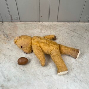 Puede incluir: Un oso de peluche vintage amarillo, de peluche, tumbado sobre una superficie de m&aacute;rmol blanco. El oso tiene la nariz marr&oacute;n y los ojos negros. Un huevo marr&oacute;n est&aacute; al lado del oso.
