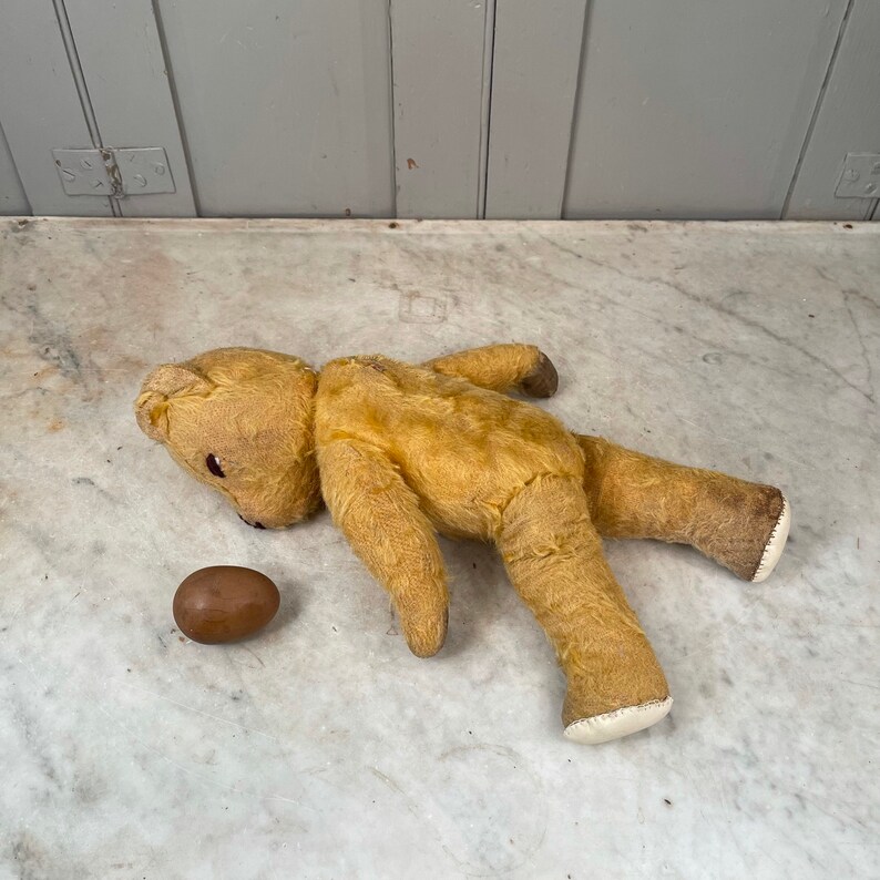 Puede incluir: Un oso de peluche vintage amarillo, de peluche, tumbado sobre una superficie de m&aacute;rmol blanco. El oso tiene la nariz marr&oacute;n y los ojos negros. Un huevo marr&oacute;n est&aacute; al lado del oso.