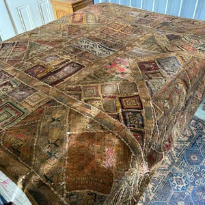 Könnte beinhalten: Eine Patchwork-Tagesdecke mit einer Vielzahl von braunen, beigen und goldenen Stoffquadraten mit komplizierten Stickmustern, geometrischen Formen und kleinen farbigen Akzenten, drapiert über einem Bettgestell mit einem weißen Metallkopfteil.