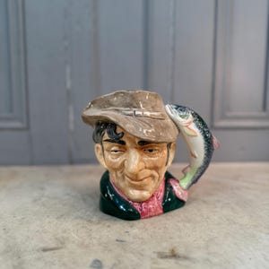 Puede incluir: Taza de cerámica con forma de cabeza de hombre, con sombrero marrón y chaqueta verde. Un pez está arqueado sobre la cabeza del hombre. La taza presenta un diseño detallado pintado a mano con una cara sonriente y una bufanda rosa.