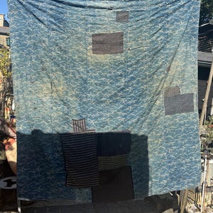 May include: A large blue textile with a white wave pattern is hanging outdoors. The textile has several dark patches and stripes sewn onto it. The textile is hanging on a washing line in front of a house and some trees.