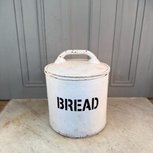 Può includere: Un contenitore per il pane bianco, cilindrico, con coperchio e manico. La parola "BREAD" è stampata in grassetto nero sul davanti. Il contenitore ha un aspetto vintage e usurato, suggerendo che si tratta di un oggetto d'antiquariato o di una riproduzione.