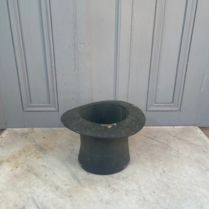 May include: A vintage black top hat with a wide brim. The hat is made of a stiff material and has a cylindrical crown. The hat is sitting on a marble surface. The hat is a classic accessory.