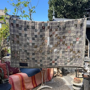 Op de afbeelding: Een vintage patchworkquilt met een ruitpatroon in tinten grijs, bruin en crème. De quilt wordt buiten tentoongesteld, hangend aan een waslijn. De quilt heeft een rasterachtig ontwerp met verschillende stoffen vierkanten.