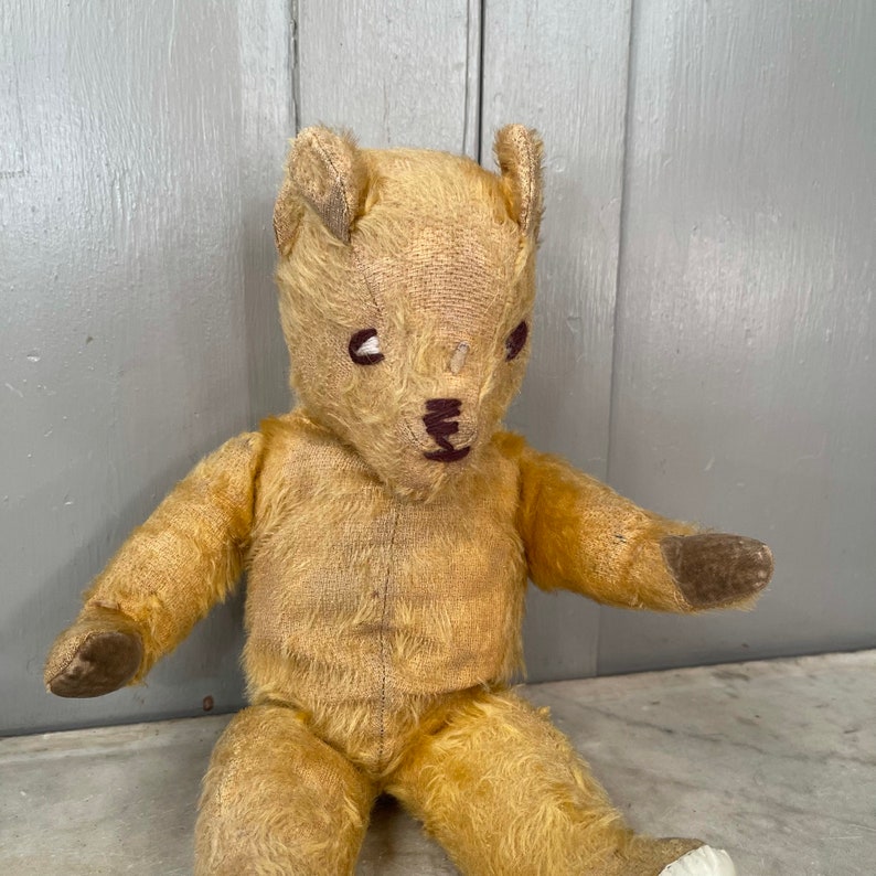 Puede incluir: Un oso de peluche vintage de color beige con ojos y nariz bordados en marr&oacute;n. El oso est&aacute; sentado con los brazos extendidos y tiene un aspecto desgastado y muy querido.