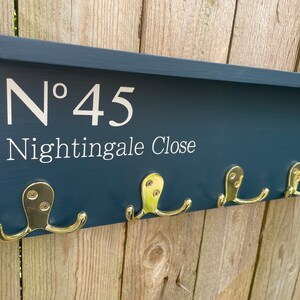 Op de afbeelding: Een donkerblauwe houten kapstok met een plank en gouden haken. De kapstok heeft het nummer "N&deg; 45 Nightingale Close" erop.