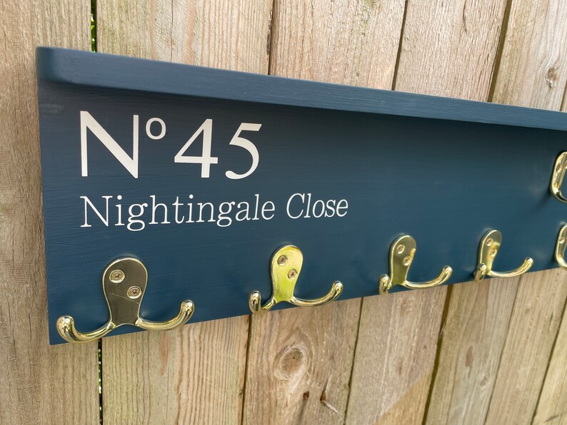 Op de afbeelding: Een donkerblauwe houten kapstok met een plank en gouden haken. De kapstok heeft het nummer "N&deg; 45 Nightingale Close" erop.