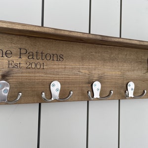 Op de afbeelding: Een houten kapstok met de tekst "The Pattons Est 2001" en zes zilveren haken.