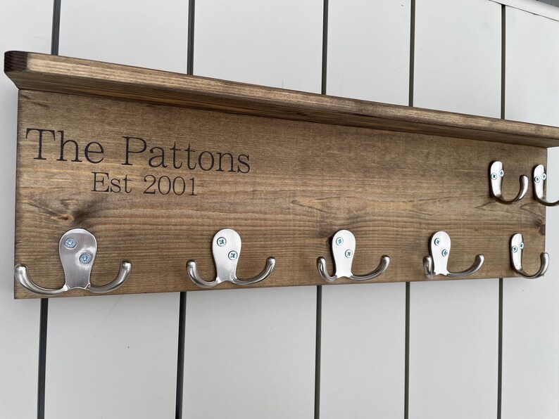Op de afbeelding: Een houten kapstok met de tekst "The Pattons Est 2001" en zes zilveren haken.