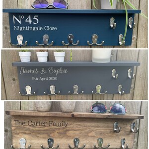 Op de afbeelding: Drie houten sleutelrekken met haken en gepersonaliseerde tekst. Het bovenste rek is donkerblauw geverfd en heeft de tekst "N&deg;45 Nightingale Close". Het middelste rek is grijs geverfd en heeft de tekst "James & Sophie 9th April 2020". Het onderste rek is bruin gebeitst en heeft de tekst "The Carter Family".