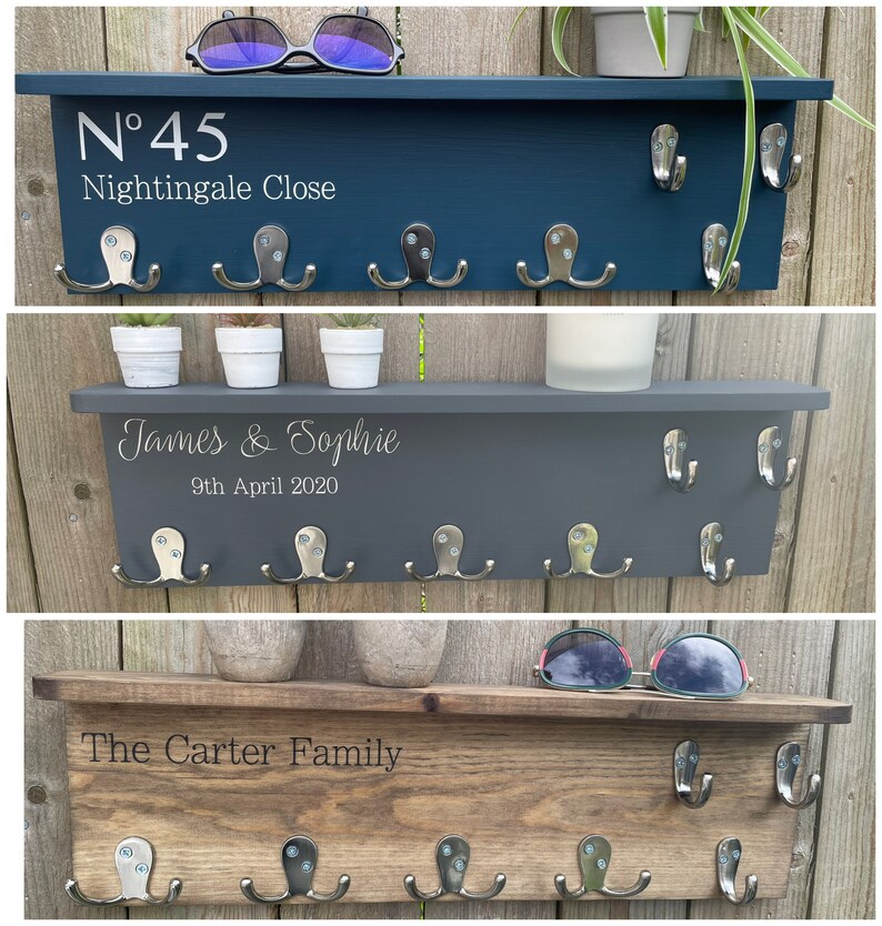 Op de afbeelding: Drie houten sleutelrekken met haken en gepersonaliseerde tekst. Het bovenste rek is donkerblauw geverfd en heeft de tekst "N&deg;45 Nightingale Close". Het middelste rek is grijs geverfd en heeft de tekst "James & Sophie 9th April 2020". Het onderste rek is bruin gebeitst en heeft de tekst "The Carter Family".