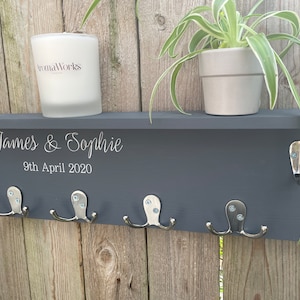 Op de afbeelding: Een grijs houten plank met vijf zilveren haken en een witte kaars. De plank heeft de namen "James & Sophie" en de datum "9 april 2020" erop gegraveerd. Een plant in een pot staat op de plank.