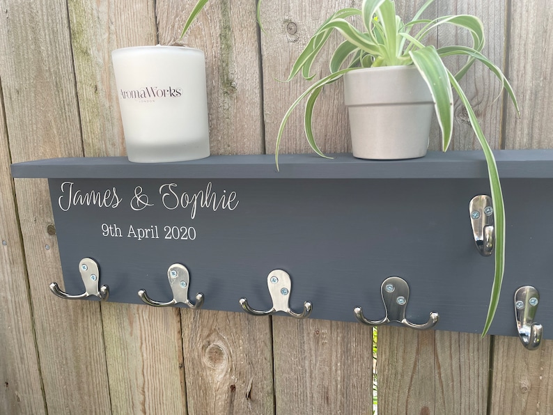 Op de afbeelding: Een grijs houten plank met vijf zilveren haken en een witte kaars. De plank heeft de namen "James & Sophie" en de datum "9 april 2020" erop gegraveerd. Een plant in een pot staat op de plank.