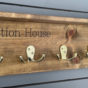 Op de afbeelding: Een houten kapstok met een plank en vijf gouden haken. De kapstok is donkerbruin gebeitst en heeft de woorden "Station House" gegraveerd op de voorkant.