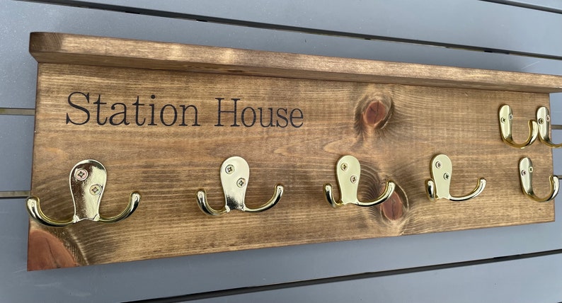 Op de afbeelding: Een houten kapstok met een plank en vijf gouden haken. De kapstok is donkerbruin gebeitst en heeft de woorden "Station House" gegraveerd op de voorkant.