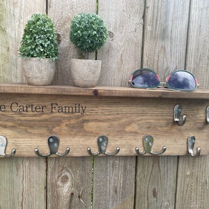 Op de afbeelding: Houten sleutelhanger met de tekst "The Carter Family" gegraveerd op de bovenste plank. De sleutelhanger heeft zes haken en is gemonteerd op een houten muur. Er staan twee kleine potplanten op de plank.