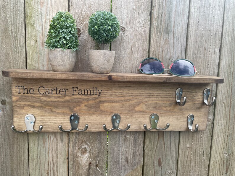 Op de afbeelding: Houten sleutelhanger met de tekst "The Carter Family" gegraveerd op de bovenste plank. De sleutelhanger heeft zes haken en is gemonteerd op een houten muur. Er staan twee kleine potplanten op de plank.