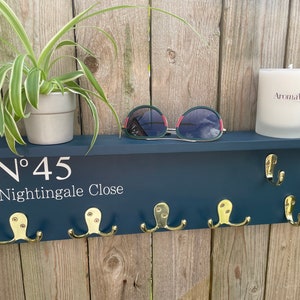 Op de afbeelding: Een donkerblauwe houten sleutelhanger met gouden haken en de tekst "N&deg; 45 Nightingale Close". Een plant in een pot, een zonnebril en een kaars staan op het plankje boven de haken.