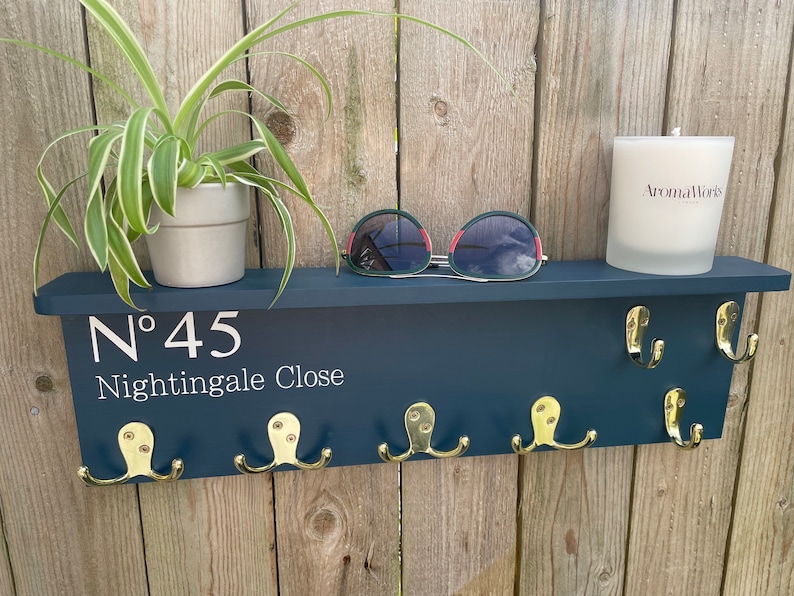Op de afbeelding: Een donkerblauwe houten sleutelhanger met gouden haken en de tekst "N&deg; 45 Nightingale Close". Een plant in een pot, een zonnebril en een kaars staan op het plankje boven de haken.