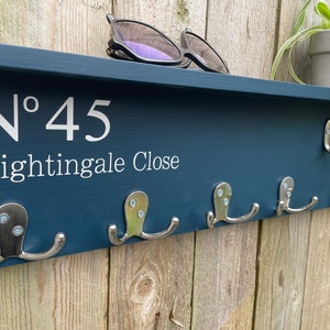 Op de afbeelding: Een donkerblauwe houten kapstok met het nummer "N&deg; 45 Nightingale Close" in wit. De kapstok heeft zes zilveren haken en een plank bovenop.