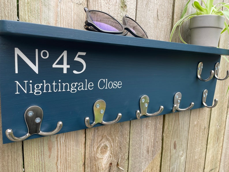 Op de afbeelding: Een donkerblauwe houten kapstok met het nummer "N&deg; 45 Nightingale Close" in wit. De kapstok heeft zes zilveren haken en een plank bovenop.