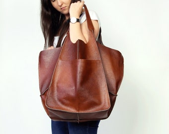 Pode incluir: Uma mulher está segurando uma grande bolsa tote de couro marrom com um único bolso na frente. Ela está vestindo jeans azul e tênis branco.