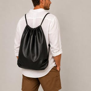 May include: A black drawstring backpack with a smooth, matte finish and thin black cord straps. The bag features a gathered top closure and a simple, unstructured design that hangs against the back, providing a clean and functional aesthetic.