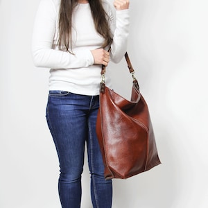Peut inclure: Une femme portant une chemise blanche &agrave; manches longues, un jean bleu et des baskets blanches tient un grand sac fourre-tout en cuir marron.