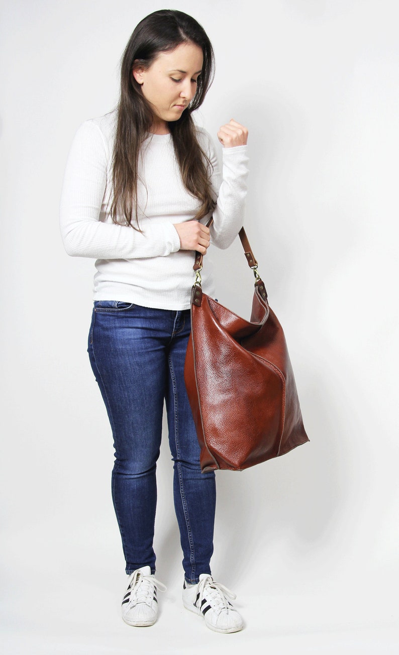 Peut inclure: Une femme portant une chemise blanche &agrave; manches longues, un jean bleu et des baskets blanches tient un grand sac fourre-tout en cuir marron.