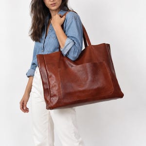 May include: A large, rectangular tote bag crafted from smooth, rich brown leather. The bag features two sturdy shoulder straps and a prominent, wide exterior pocket centered on the front panel. The leather has a natural, slightly textured finish.