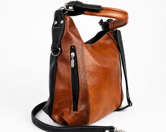 May include: A two-tone hobo bag featuring a textured cognac brown center panel and smooth black side panels. The bag includes a vertical zippered pocket on the front, a top handle, and a detachable black shoulder strap with silver-toned hardware.