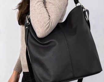 Op de afbeelding: Een zwarte leren schoudertas met een lange, verstelbare riem. De tas heeft een eenvoudig, minimalistisch ontwerp met een groot hoofdvak en een kleinere riem om te dragen. De tas wordt getoond tegen een neutrale achtergrond.