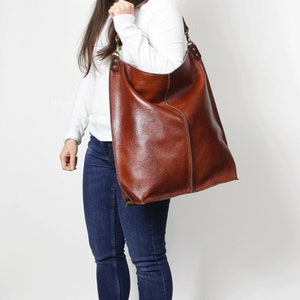 Peut inclure: Un sac cabas en cuir marron avec une longue bandouli&egrave;re. Le sac est port&eacute; par une personne v&ecirc;tue d'une chemise blanche et d'un jean bleu.