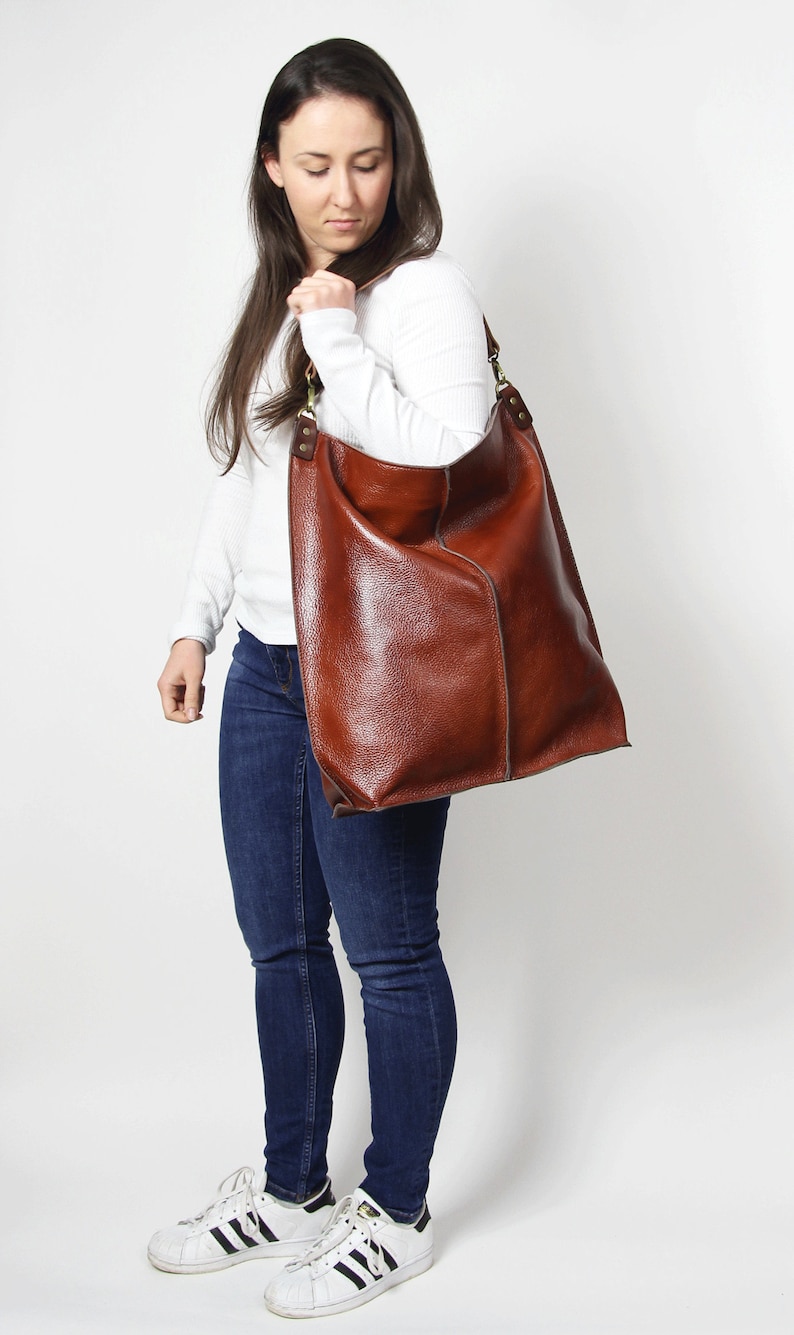 Peut inclure: Un sac cabas en cuir marron avec une longue bandouli&egrave;re. Le sac est port&eacute; par une personne v&ecirc;tue d'une chemise blanche et d'un jean bleu.