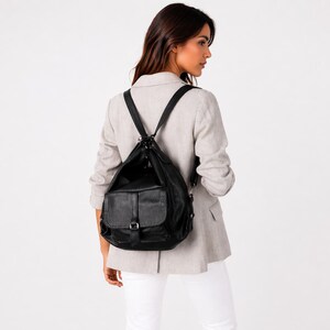 Peut inclure: Un sac à dos en cuir noir avec une poche à rabat et des bretelles réglables. Le sac à dos est porté sur les épaules. La personne porte un blazer gris clair et un pantalon blanc.