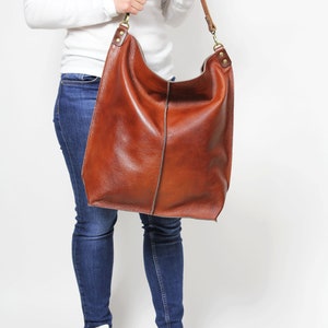 Peut inclure: Un grand sac fourre-tout en cuir marron avec une longue bandouli&egrave;re. Le sac est port&eacute; par une personne v&ecirc;tue d'un jean bleu et de baskets blanches.